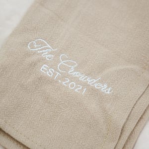 Pode incluir: Tecido de linho bege com texto bordado branco: "The Crowders EST. 2021". O tecido está dobrado, mostrando uma superfície texturizada. O bordado é em letra cursiva.