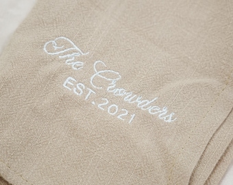 Puede incluir: Tela de lino beige con texto bordado en blanco: "The Crowders EST. 2021". La tela está doblada, mostrando una superficie texturizada. El bordado está en letra cursiva.