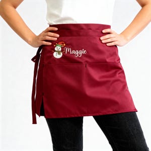 May include: A burgundy waist apron with two pockets and side ties. The apron features an embroidered snowman wearing a hat and scarf, with the name "Maggie" embroidered in cursive script.