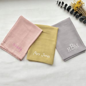 Pode incluir: Três guardanapos de linho dobrados em rosa, dourado e cinza, cada um bordado com texto personalizado. O guardanapo rosa diz "Wendy", o dourado "Mrs Jones" e o cinza tem as iniciais "HBM".