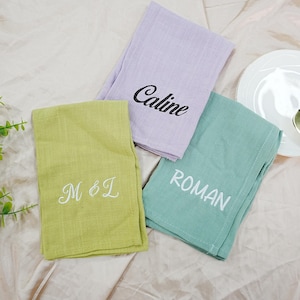May include: Three folded linen towels in pastel shades: light green, lavender, and teal. Each towel is embroidered with a name or initials in white thread: "M & L", "Caline", and "Roman".