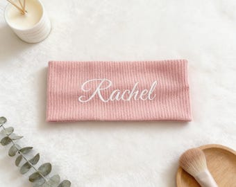Puede incluir: Una diadema rosa con el nombre "Rachel" bordado en blanco. La diadema tiene una textura acanalada y se muestra sobre una superficie blanca texturizada. Una vela y una brocha de maquillaje son visibles en el fondo.