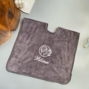Puede incluir: Una toalla de spa gris con un corte para el cuello. Tiene un diseño bordado en blanco del perfil de una mujer, una flor y el nombre "Melanie". La toalla está hecha de un material suave y afelpado.
