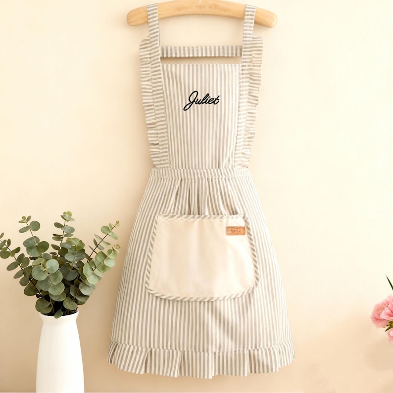 May include: A striped apron with the name "Juliet" embroidered on the front. The apron has a large pocket and ruffled edges. It is hanging on a wooden hanger.
