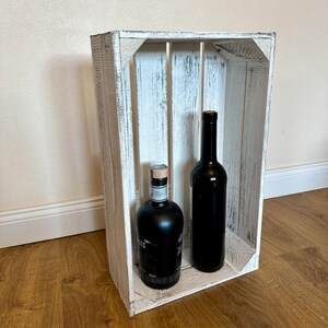 Könnte beinhalten: Weiße Holzkiste mit zwei Flaschen Spirituosen darin. Die Kiste hat eine weiße, abgenutzte Lackierung. Die Flaschen sind beide aus dunklem Glas und haben Etiketten.