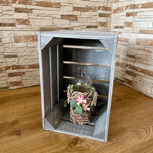 Könnte beinhalten: Eine graue Holzkiste mit einer abgenutzten Oberfläche. Die Kiste hat eine Glasflasche im Inneren mit einem Weidenkorb gefüllt mit rosa und grünen Kunstblumen.