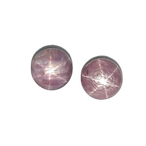 Könnte beinhalten: Zwei rosa Stern-Saphire mit einem sechszackigen Sternmuster. Die Saphire sind rund und im Cabochon-Schliff.