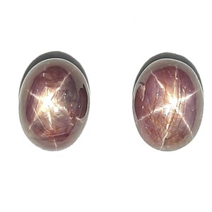 Könnte beinhalten: Zwei ovale, rote Stern-Saphire mit einem sechszackigen Stern in der Mitte. Die Steine sind im Cabochon-Schliff geschliffen und haben eine rotbraune Farbe.