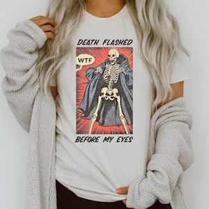 Può includere: Una maglietta bianca con una grafica in stile fumetto vintage di uno scheletro in una veste nera. Il testo sulla maglietta recita "Death Flashed WTF Before My Eyes".
