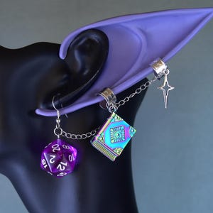 Könnte beinhalten: Fantasy-Ohrringe mit einem lila D20-Würfel und einem farbwechselnden Buchanhänger. Die Ohrringe sind durch eine Kette verbunden und an einer silbernen Ohrklemme befestigt. Ein lila Elfenohr ist ebenfalls sichtbar.