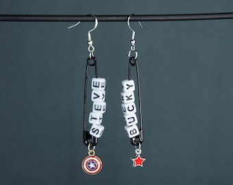 May include: A pair of handmade earrings featuring black safety pins. One earring spells "STEVE" with white letter beads and has a shield charm. The other spells "BUCKY" and has a red star charm. Silver-colored ear wires.