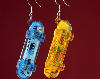 May include: A pair of dangle earrings featuring miniature skateboard designs. One earring is blue with a white graphic and the other is yellow with a red, orange, and yellow graphic.