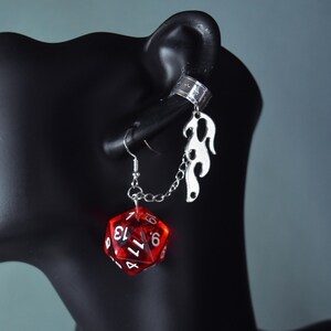 May include: A distinctive earring set with a red, multi-sided die displaying white numbers, linked by a chain to a silver flame ear cuff. The earring is shown on a black mannequin head against a grey backdrop.