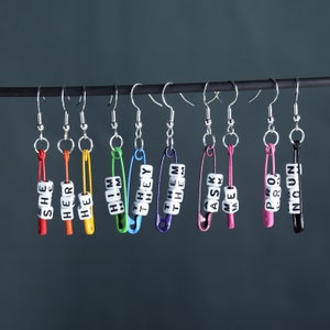 Può includere: Un set di 10 orecchini a forma di spilla di sicurezza colorati con diverse parole di pronomi scritte su ciascuno. Gli orecchini sono appesi a una barra argentata.