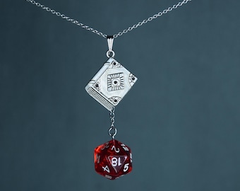 May include: A silver pendant necklace with a book-shaped charm and a red, twenty-sided die. The book charm has etched details. The die is suspended below the book charm by a small chain. The necklace chain is silver-toned.