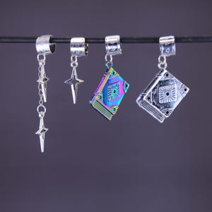 Può includere: Perline a forma di ciondolo argentate con disegni di stelle e libri. Un ciondolo presenta una catena di tre stelle, mentre altri presentano forme di stelle e libri aperti. I ciondoli a forma di libro hanno finiture iridescenti e argentate.