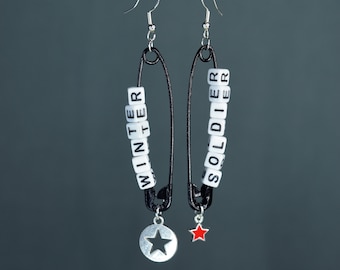 May include: A pair of black safety pin earrings with white letter beads spelling "WINTER SOLDIER" and star charms. The left earring has a silver star charm and the right earring has a red star charm.
