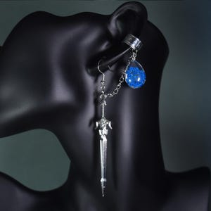 Op de afbeelding: Een zilverkleurige oormanchet- en oorbellen set. De oorbel heeft een zwaardhanger en een druppelvormige bedel gevuld met blauwe glitter. Een ketting verbindt de oorbel met de oormanchet. De sieraden worden getoond op een zwarte mannequin hoofd.