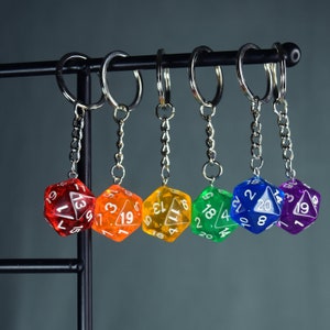 Könnte beinhalten: Sechs bunte, transparente Plastikwürfel mit weißen Zahlen auf jeder Seite, die an silbernen Schlüsselanhängern an einem schwarzen Metallgestell hängen. Die Würfel sind rot, orange, gelb, grün, blau und lila.