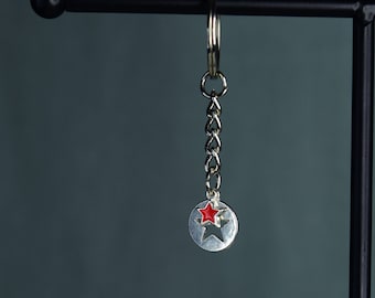 May include: A silver keychain with a red star charm. The star is in the center of a circle with a star cut out of it.