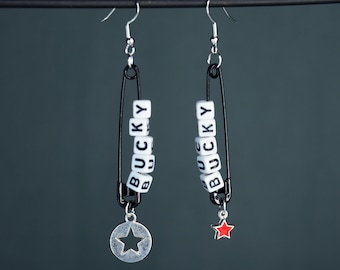 May include: A pair of silver earrings with black safety pins and white letter beads spelling "BUCKY". One earring has a silver star charm, the other has a red star charm.