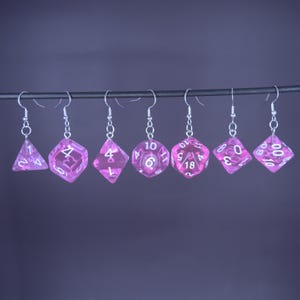 Può includere: Un set di sette orecchini a forma di dado rosa, ciascuno con numeri bianchi e ganci argentati. I dadi includono un triangolo, un quadrato e vari poliedri. Sfondo viola.