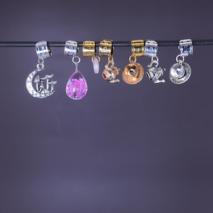 May include: A selection of decorative charms, including a silver crescent moon with mushrooms, a pink teardrop with flowers, and gold and silver teapot and teacup designs. The charms are suspended from a dark rod against a purple backdrop.