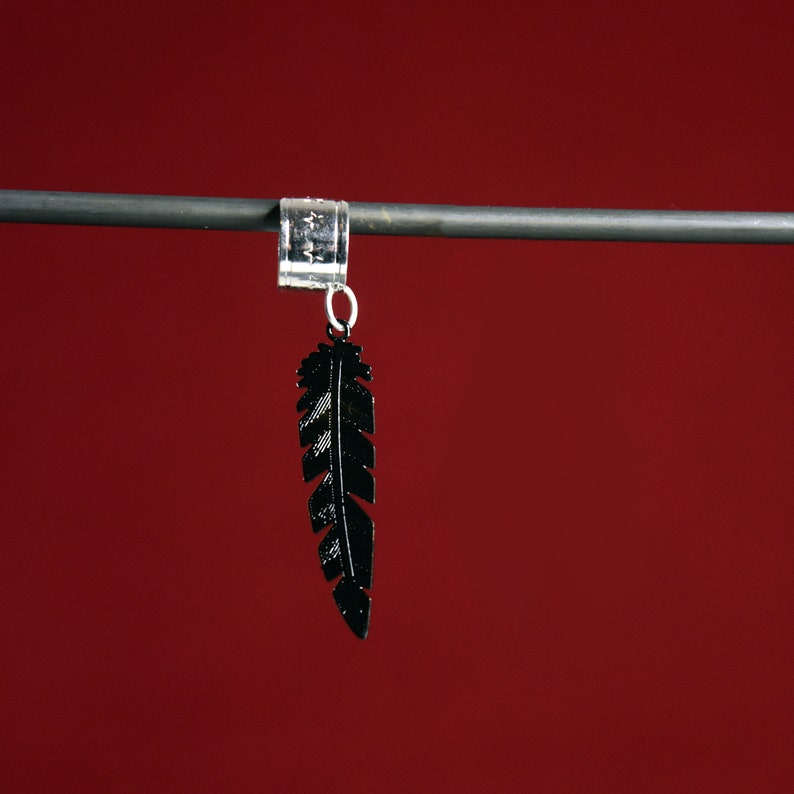 May include: A black feather charm hangs from a silver ring on a metal rod. The feather is detailed and has a slightly textured surface.
