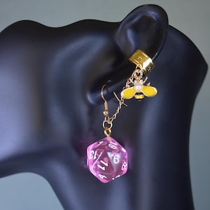 May include: An unusual earring set with a gold-tone ear cuff and a dangle earring. The dangle earring has a pink, transparent, twenty-sided die and a gold chain with a yellow and black bee charm. The ear cuff has a gold star design.