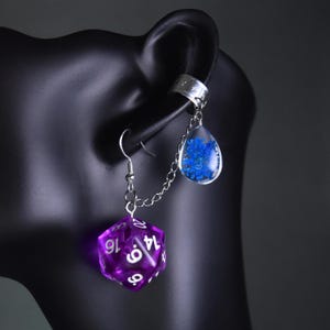 May include: A distinctive earring set with a purple, glowing, twenty-sided die and a teardrop-shaped charm filled with blue flowers. The earring is connected to a silver ear cuff with a delicate chain.