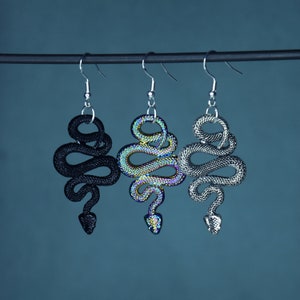Könnte beinhalten: Drei schlangenförmige Ohrringe mit silbernen Haken. Einer ist schwarz, einer hat ein schimmerndes, irisierendes Finish und der dritte ist silberfarben. Die Ohrringe hängen vor einem teal-farbenen Hintergrund.