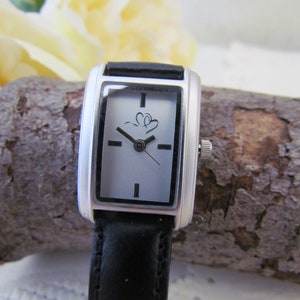 Può includere: Un orologio da polso rettangolare argento e nero con quadrante bianco e cinturino in pelle nera. Il quadrante dell'orologio ha due lancette e un piccolo logo con due cuori.