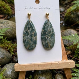 Puede incluir: Un par de pendientes en forma de lágrima con un patrón de mármol verde oscuro y motas doradas. Los pendientes están sujetos a una tarjeta blanca con la inscripción Fashion Jewelry y tienen postes de metal dorado.