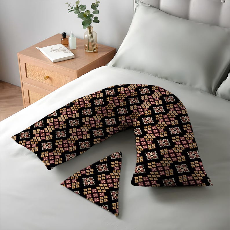 K&ouml;nnte beinhalten: Ein schwarzes V-f&ouml;rmiges K&ouml;rperkissen und ein dreieckiges Kissen mit einem sich wiederholenden Blumenmuster in Beige und Rosa. Die Kissen liegen auf einem wei&szlig;en Bett.