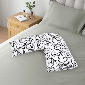 K&ouml;nnte beinhalten: Ein V-f&ouml;rmiges Kissen mit einem schwarz-wei&szlig;en Blumenmuster auf einem grauen Bett. Das Kissen hat ein sich wiederholendes Muster aus schwarzen Wirbeln und Bl&auml;ttern. Ein Nachttisch aus Holz mit einem Buch und einer Vase befindet sich im Hintergrund.