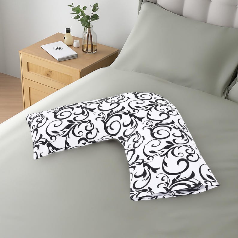 K&ouml;nnte beinhalten: Ein V-f&ouml;rmiges Kissen mit einem schwarz-wei&szlig;en Blumenmuster auf einem grauen Bett. Das Kissen hat ein sich wiederholendes Muster aus schwarzen Wirbeln und Bl&auml;ttern. Ein Nachttisch aus Holz mit einem Buch und einer Vase befindet sich im Hintergrund.