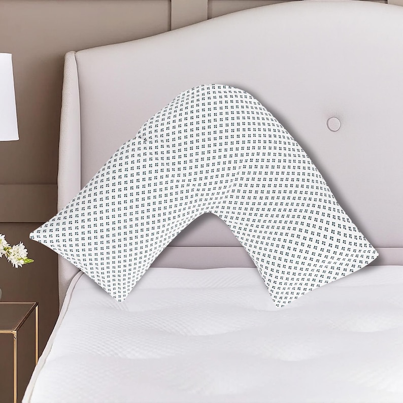 K&ouml;nnte beinhalten: Ein wei&szlig;es, V-f&ouml;rmiges Kissen mit einem sich wiederholenden Muster aus kleinen, dunkelblauen floralen Designs. Das Kissen liegt auf einem wei&szlig;en Bett, mit einem hellrosa Kopfteil im Hintergrund. Das Kissen ist f&uuml;r Komfort und Unterst&uuml;tzung konzipiert.