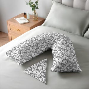 K&ouml;nnte beinhalten: Ein V-f&ouml;rmiges Kissen und ein kleines dreieckiges Kissen mit einem wei&szlig;en und grauen Scrollmuster auf einem Bett. Die Bettw&auml;sche ist hellgrau. Die Kissen sind f&uuml;r Komfort und Unterst&uuml;tzung konzipiert.
