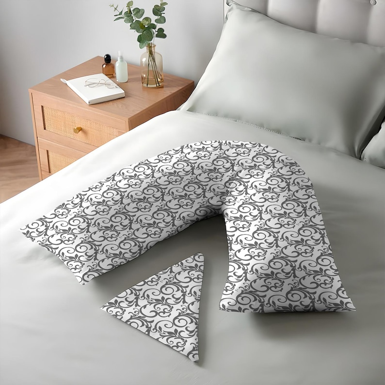 K&ouml;nnte beinhalten: Ein V-f&ouml;rmiges Kissen und ein kleines dreieckiges Kissen mit einem wei&szlig;en und grauen Scrollmuster auf einem Bett. Die Bettw&auml;sche ist hellgrau. Die Kissen sind f&uuml;r Komfort und Unterst&uuml;tzung konzipiert.