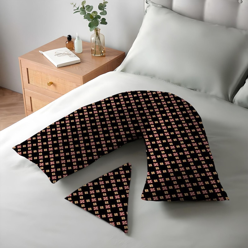 K&ouml;nnte beinhalten: Ein schwarzes K&ouml;rperkissen und ein kleines dreieckiges Kissen mit einem sich wiederholenden Blumenmuster in Rot und Creme. Die Kissen liegen auf einem wei&szlig;en Bett. Ein Nachttisch ist im Hintergrund.