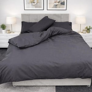 May include: A charcoal grey bed set, including a duvet cover and pillowcases, on a bed with a grey headboard. The bedding is neatly arranged, creating a clean look. Two white bedside tables with lamps and decor are on either side of the bed.