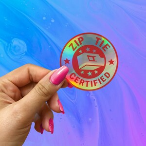 May include: A holographic sticker with a red circle and the text "ZIP TIE CERTIFIED" in white. The circle has a white outline and five white stars around the edge. Inside the circle is a white outline of a zip tie.