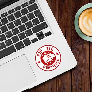 May include: A red and white sticker on a silver laptop. The sticker is a circle with the words "ZIP TIE CERTIFIED" and a graphic of a zip tie.