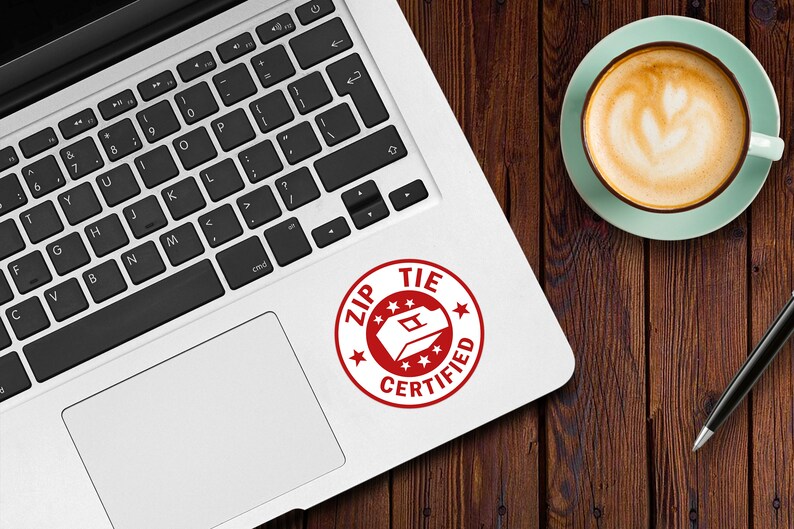 May include: A red and white sticker on a silver laptop. The sticker is a circle with the words "ZIP TIE CERTIFIED" and a graphic of a zip tie.