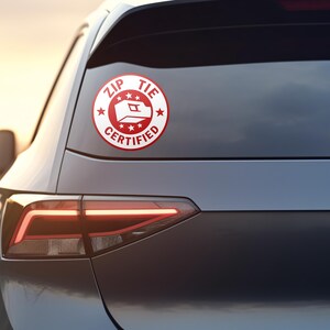May include: A red and white sticker with the text "ZIP TIE CERTIFIED" and a stylized image of a zip tie. The sticker is on the back window of a gray car.