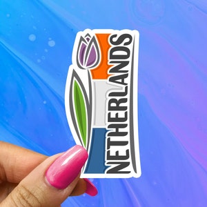 May include: A sticker featuring the Netherlands flag design with a purple tulip and the word "NETHERLANDS" in white. The flag is orange, white, and blue. The sticker is held against a blue and purple background.