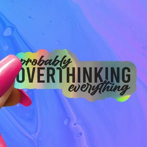 May include: A holographic sticker with the text "probably overthinking everything" in black lettering. The sticker is being held by a hand with pink nail polish.