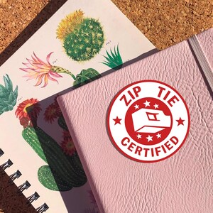 May include: A pink notebook with a red and white sticker that says "Zip Tie Certified" with a white star design.