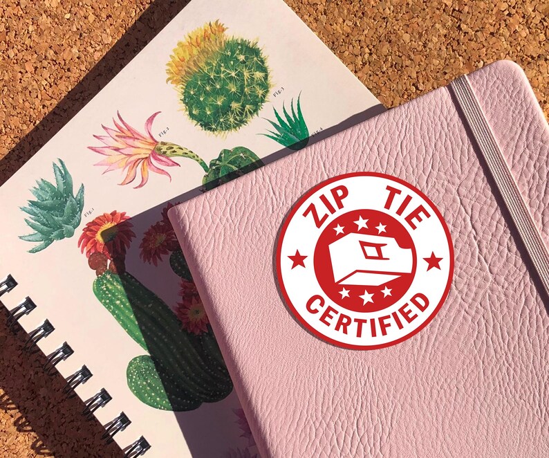 May include: A pink notebook with a red and white sticker that says "Zip Tie Certified" with a white star design.