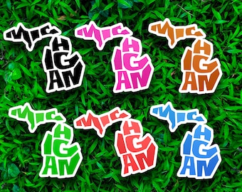 May include: Six Michigan state outline stickers in different colors: black, pink, orange, green, red, and blue. Each sticker has the word "MICHIGAN" spelled out within the state outline.